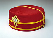 KCCH Cap
The distinctive regalia of a Knights Commander of the Court of Honour is the red cap.  Made of rich fabric, the cap is red with a dark red band trimmed in gold.  It has a gold cord which extends across the top.  In the center front is a representation of the Knight Commander jewel, a Passion Cross with fancy arms, featuring in the center a trefoil embroidered in a green encircled by “Kt. Commander Court of Honour” embroidered in gold.